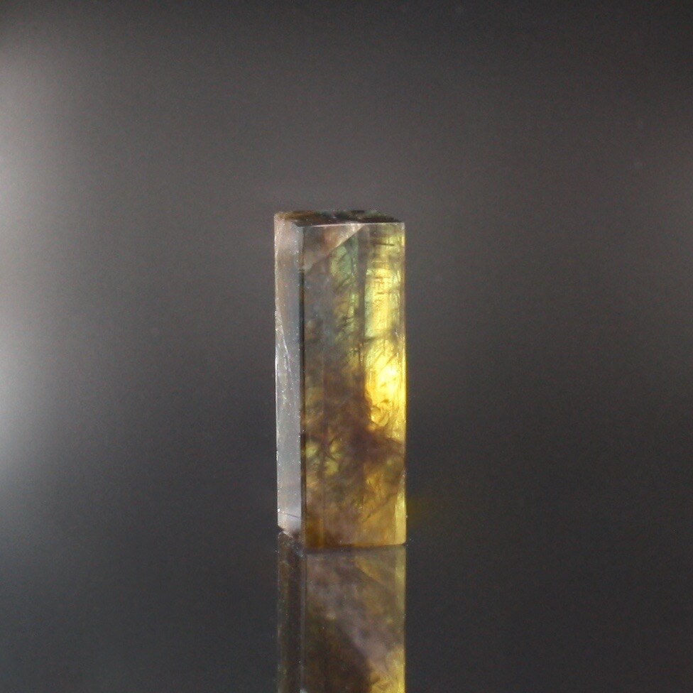 Elongated rectangular step-cut gold sheen sapphire in yellow and green with dark hair-like inclusions, against a dark background.