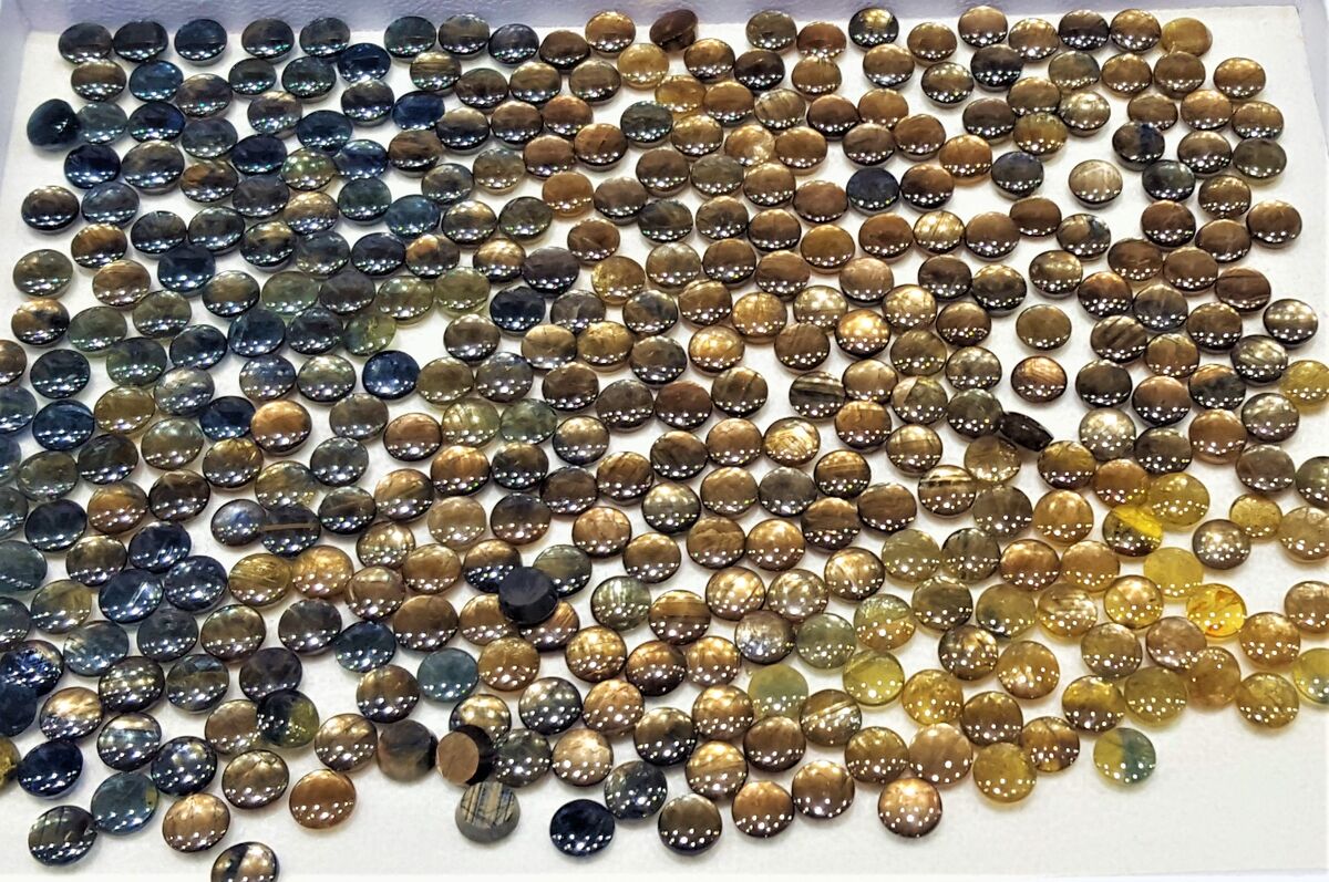 Several hundred round gold sheen sapphire cabochons spread across a white tray, shading from blue and grey on the left through gold and brown to yellow on the right.