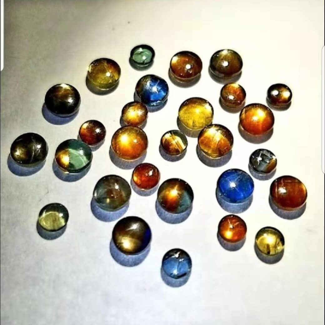 About thirty round gold sheen sapphire cabochons scattered on a pale grey surface, in blue, green, yellow, orange and brown, many with six-rayed stars.