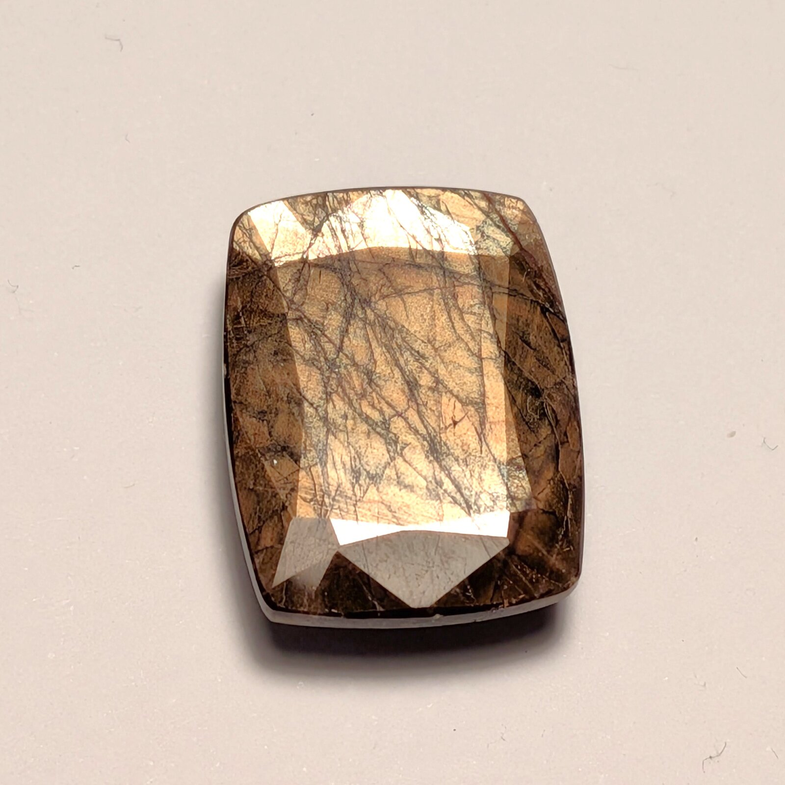 Rectangular cushion-shaped faceted brown gem with a bright golden sheen across its table, crossed by a web of fine dark lines, on a pale grey background.