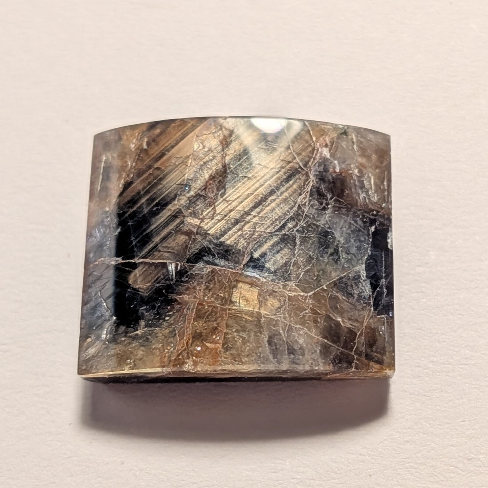 Rectangular gently domed gem in mottled blue-black and brown, with parallel golden streaks running diagonally across it and a network of fine pale lines, on a pale grey background.